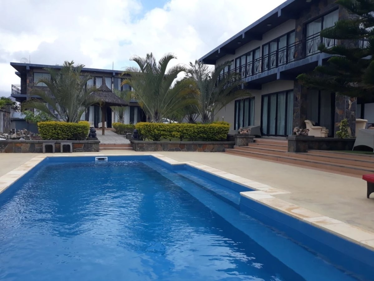A large outdoor swimming pool occupies the foreground, with steps leading into the water. The surrounding area features well-maintained landscaping, including palm trees and shrubs. In the background, two spacious villas offer expansive glass doors, contributing to a serene atmosphere.