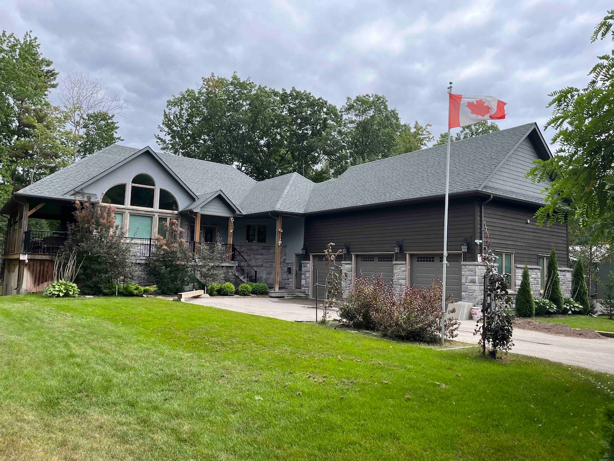 A modern bungalow is set amidst lush greenery, featuring a stone facade and a prominent red Canadian flag. A spacious driveway leads to a three-car garage. Various shrubs and landscaping elements enhance the front yard, creating an inviting entrance to the home.