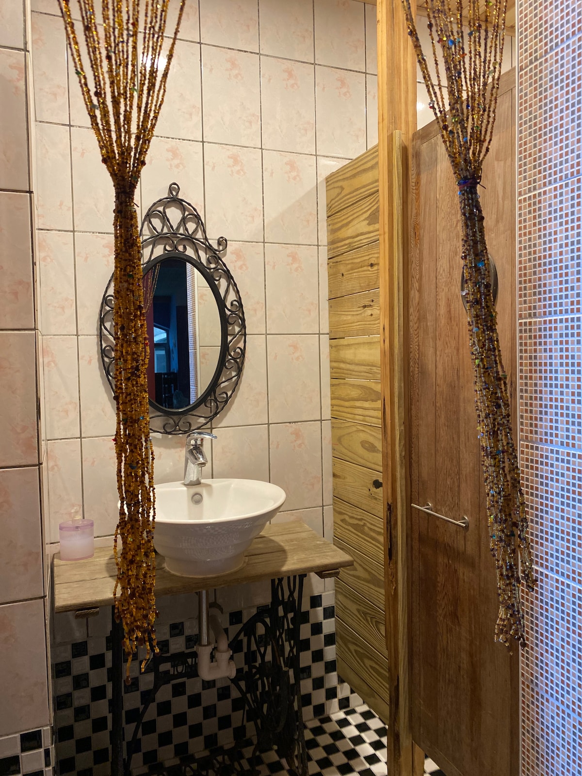 A compact bathroom area features a decorative mirror above a round sink on a wooden countertop. This space is separated by a curtain-like beaded design, with textured walls displaying a mix of tiles in light and dark hues. The flooring is a checkered pattern.