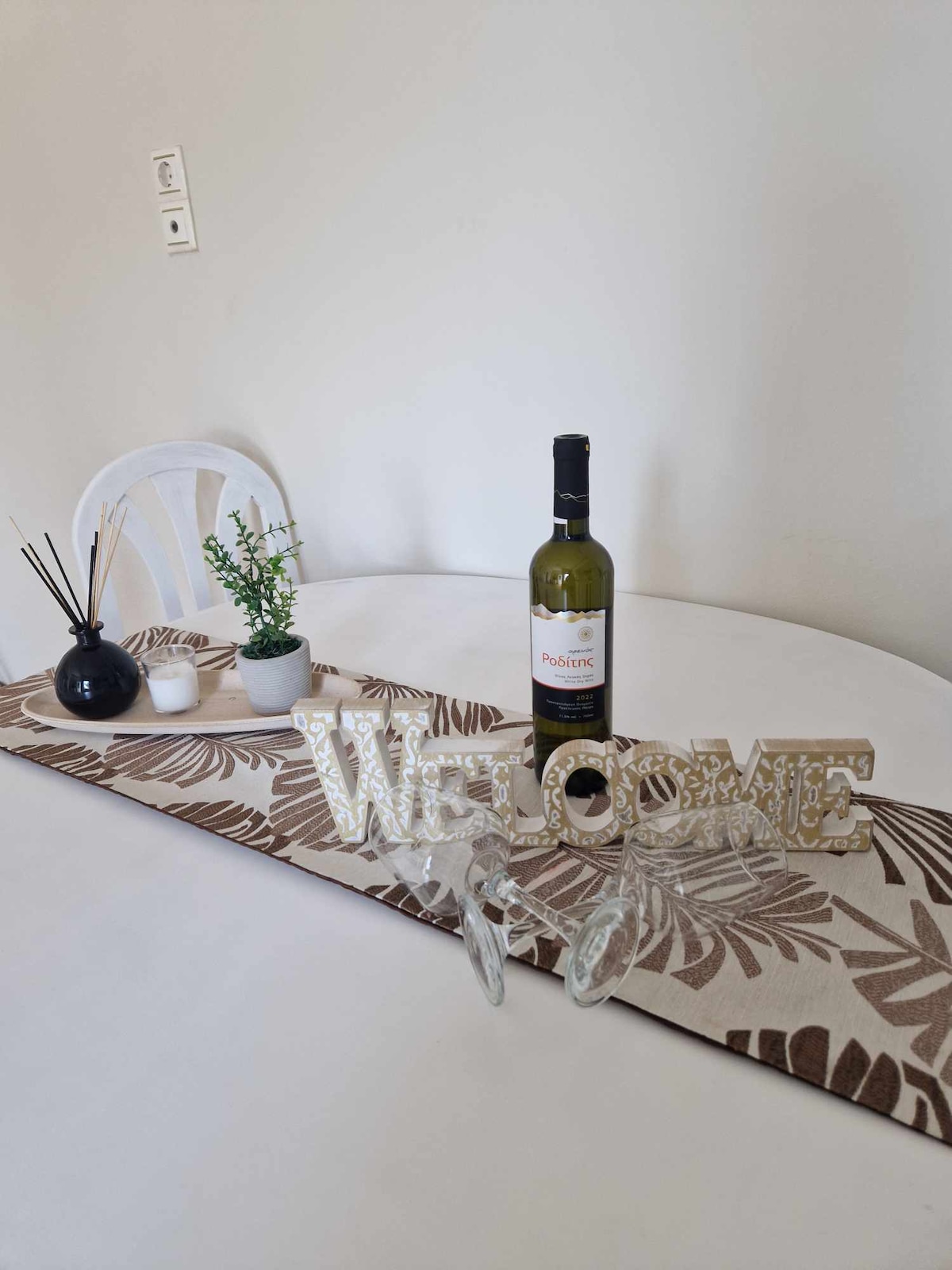 A dining table is set with a decorative runner featuring floral patterns. A bottle of wine stands at the center, accompanied by two elegant glasses. Small potted plants and a diffuser add a touch of greenery, while a 'Welcome' sign is prominently displayed.