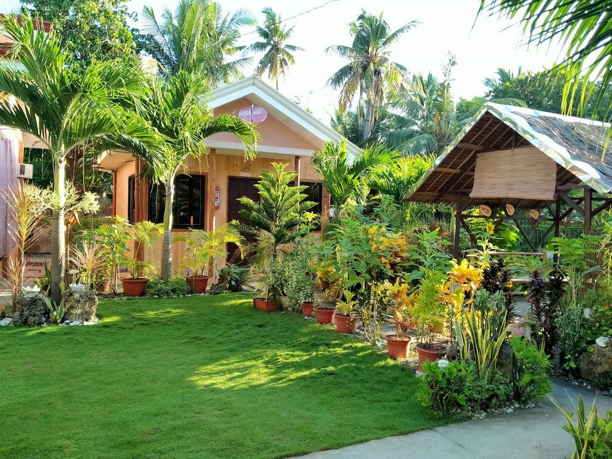 The exterior showcases a charming wooden structure surrounded by lush greenery and vibrant tropical plants. A manicured lawn compliments the setting, while a shaded outdoor dining area under a roof is visible to the side, enhancing the inviting ambience of the space.