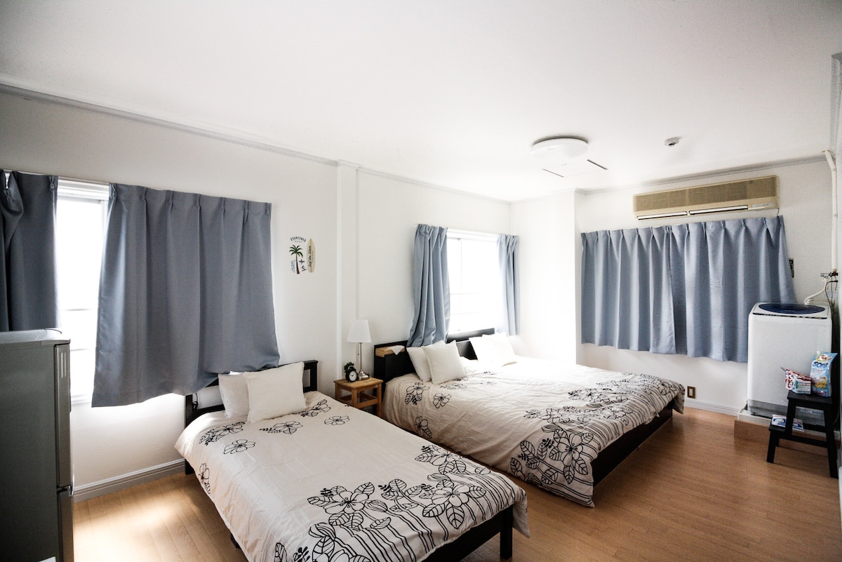 The room features two spacious beds dressed in light bedding, accompanied by decorative pillows. Natural light enters through multiple windows adorned with gray curtains. A small bedside table with a lamp stands beside the beds, while a cool air conditioning unit is installed on the wall.