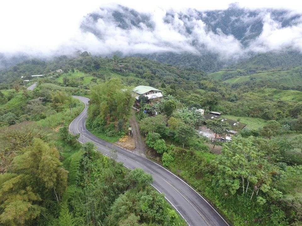 An aerial view captures a hillside retreat nestled among lush greenery, surrounded by misty mountains. The winding road leads to the property, which features a multi-story building partially obscured by trees. The natural landscape displays a vibrant mix of vegetation and rolling hills.