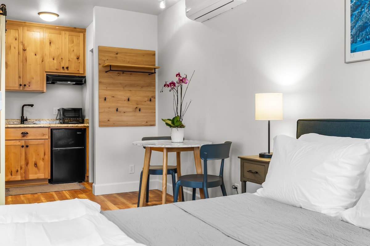 The studio features light wooden cabinetry in the kitchenette, equipped with a small refrigerator and a microwave. A round table with two chairs is positioned beside a flower arrangement, while a spacious bed is visible in the foreground, complemented by soft white linens.