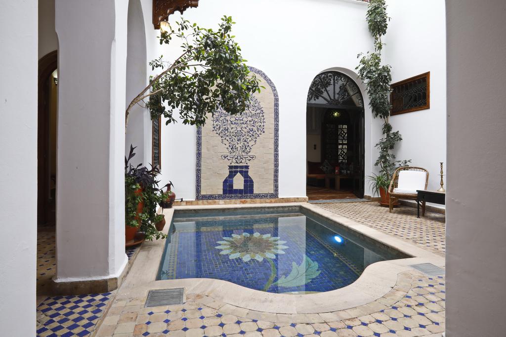 A serene courtyard is highlighted by a small, tiled pool featuring a floral design at its center. Surrounding greenery and decorative tiles create a tranquil ambiance, while white walls enhance natural light. Arched doorways lead to additional areas of the space.