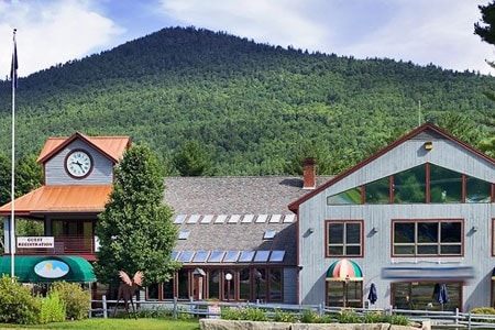 The lodge exterior is framed by a backdrop of mountains, showcasing a rustic design with a prominent clock tower. Large windows provide ample natural light, while trees and colorful awnings enhance the inviting ambiance of the building.