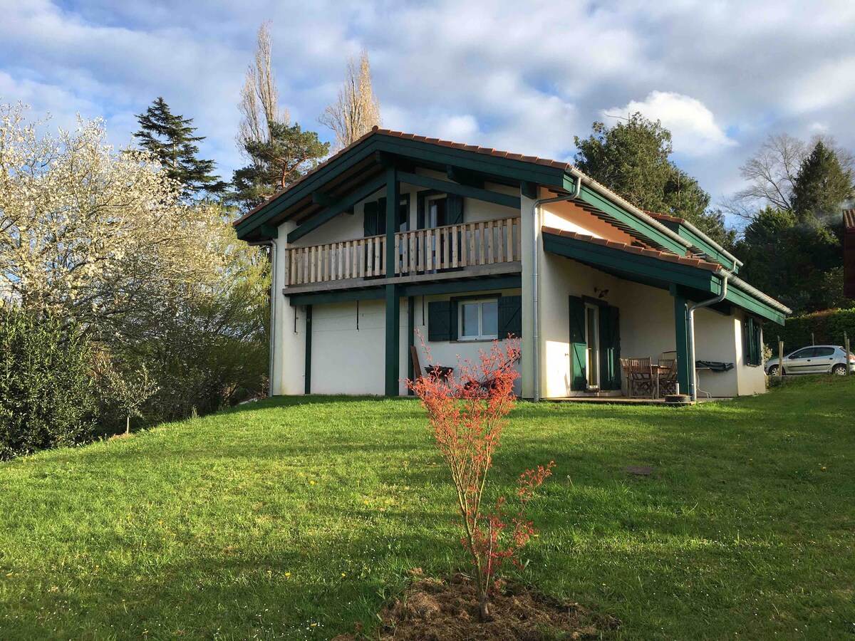 A charming two-story wooden house is set amidst lush greenery. The exterior features a balcony with wooden railing, large windows, and a door opening to a terrace. A small plant is visible in the foreground, enhancing the home's serene environment.