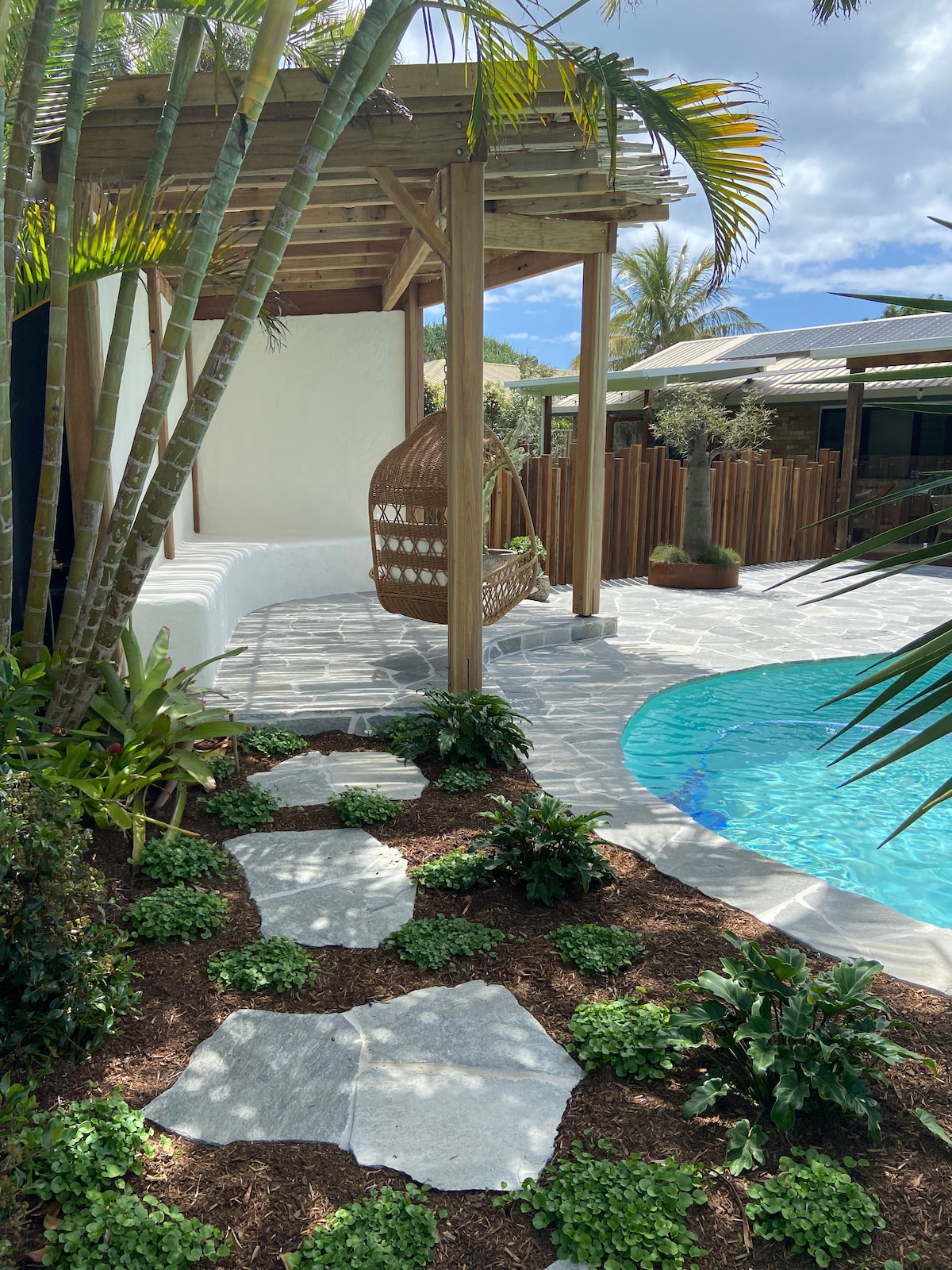 A serene outdoor space features a circular stone path leading to a sparkling pool. A wooden pergola provides shade, with a hanging wicker chair inviting relaxation. Lush greenery and low-maintenance plants surround the area, enhancing the tranquil setting.