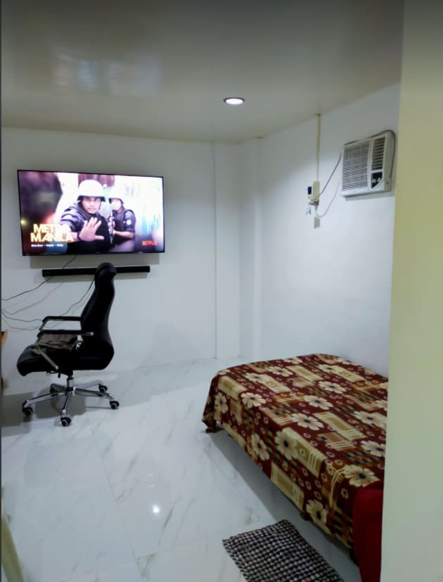 A minimalist guest suite is shown, featuring a spacious bed with patterned bedding. A wall-mounted television is positioned opposite the bed, while a desk chair is situated beside a small desk. An air conditioning unit is installed above the bed for climate control.
