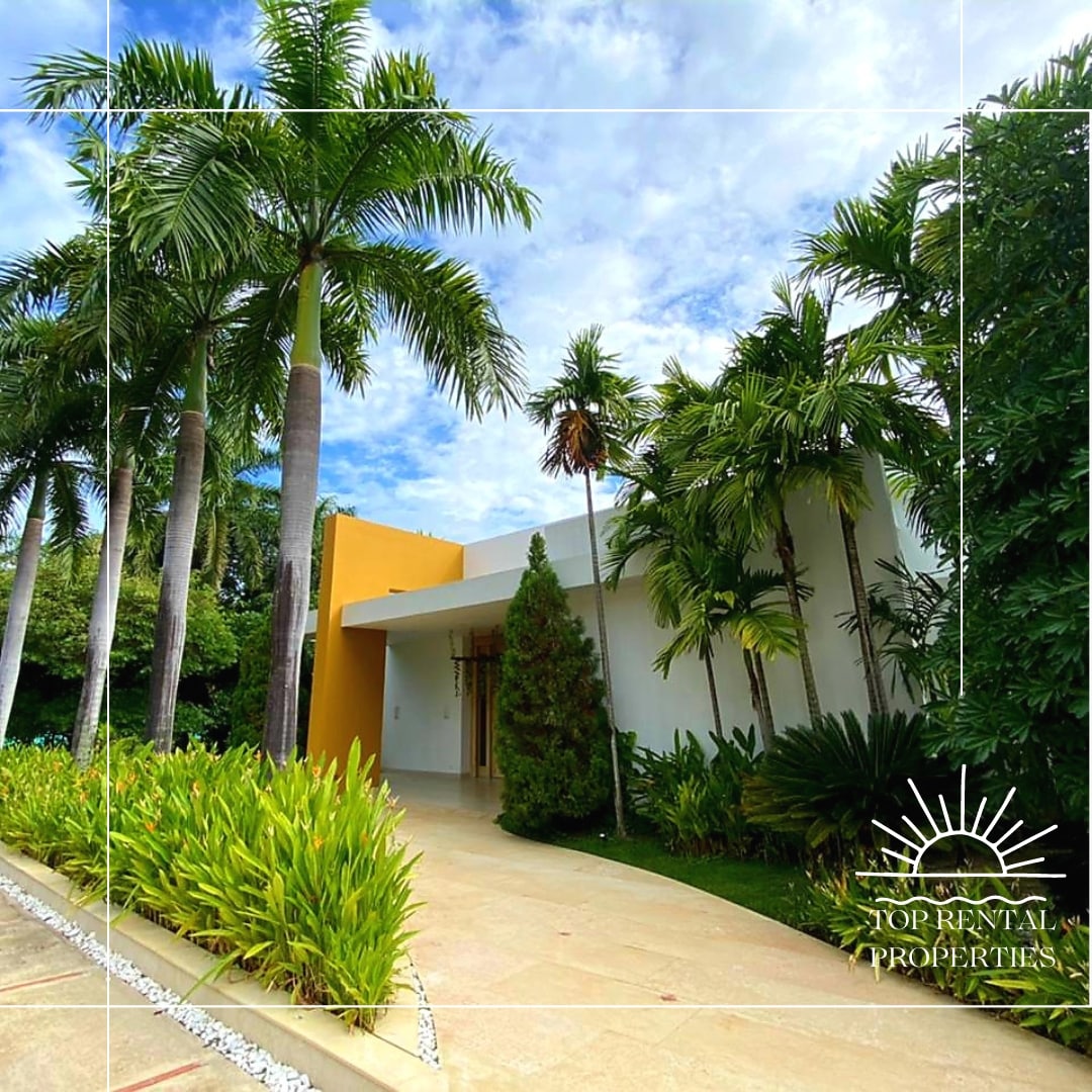 The entrance of a modern property is framed by lush green palms and neatly trimmed hedges. A smooth stone pathway leads to the white facade, featuring large windows. Bright yellow walls contrast with the surrounding greenery under a partly cloudy sky.