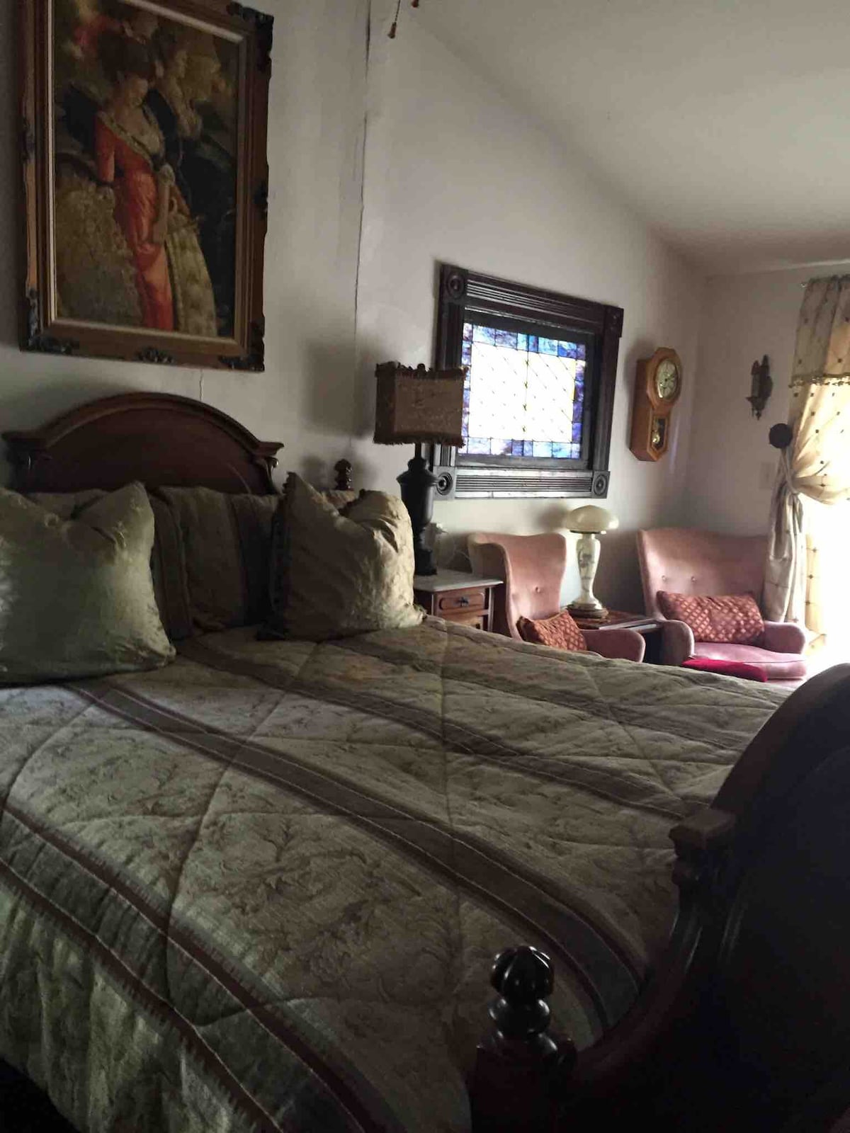A cozy sleeping area features a king size bed adorned with plush pillows and a detailed quilt. A portrait hangs above the bed, complemented by vintage lamps. A sitting area with two chairs offers a comfortable spot for relaxation, illuminated by natural light from the nearby window.