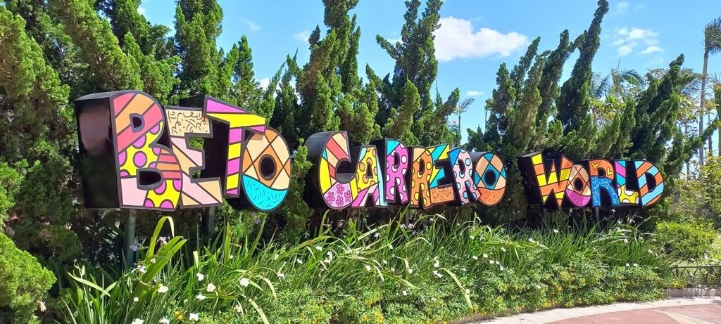 A colorful sign for Beto Carrero World is surrounded by lush green plants and vibrant flowers. The artistic letters are prominently displayed against a bright blue sky, providing an inviting entrance to the amusement park.