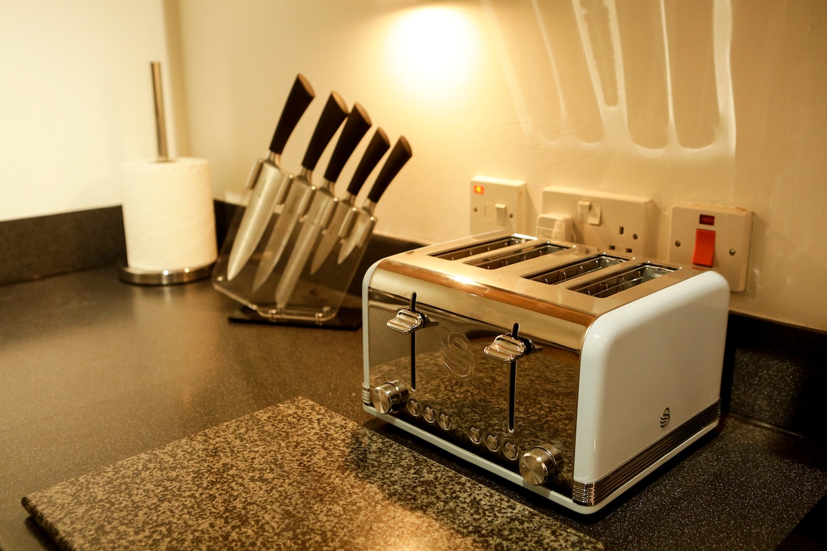 A modern kitchen countertop is displayed, featuring a sleek white toaster with four slots. Sharp knives are organized in a stand, positioned against the wall with a paper towel dispenser nearby. Soft lighting highlights the clean surfaces.