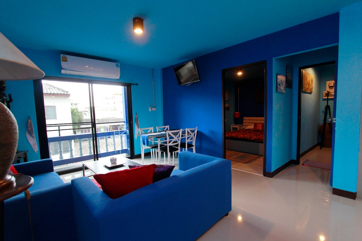 A vibrant living area features bold blue walls and bright furnishings, including two inviting sofas and a dining table for four. Large glass doors lead to a balcony, providing natural light. A television is mounted on the wall, and a doorway leads to a cozy sleeping space.