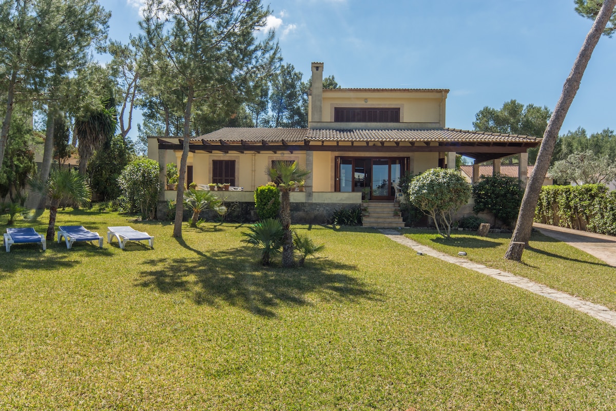 The villa is set amidst a well-maintained garden featuring lush green grass and several palm trees. A spacious terrace with outdoor seating is visible at the front, framed by the villa's two-story structure. Sun loungers are arranged on the lawn, inviting moments of relaxation.