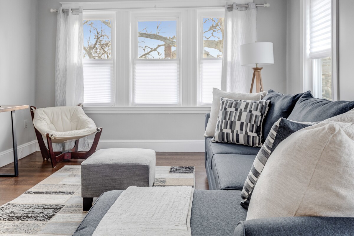 A bright living area features large windows with sheer white curtains, allowing natural light to fill the space. A comfortable sectional sofa is paired with a matching ottoman and a rounded chair, while a textured area rug lies beneath. A minimalist lamp stands in the corner.