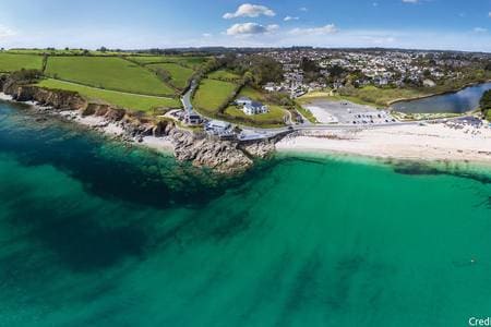 The image captures a scenic coastal view with clear turquoise waters gently lapping at a sandy beach. Lush green fields rise in the background, and a winding road leads to a nearby parking area. The town is visible in the distance, enhancing the tranquil setting.