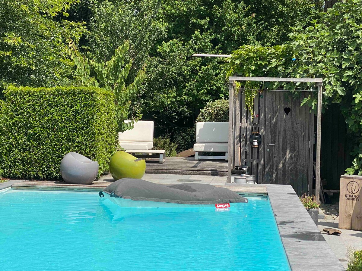 A serene swimming pool is framed by lush greenery and neatly trimmed hedges. Comfortable seating areas are positioned nearby, featuring soft seating options in varied shapes. A rustic wooden structure houses a lantern, enhancing the outdoor space's charm. Natural light reflects off the pool's surface.