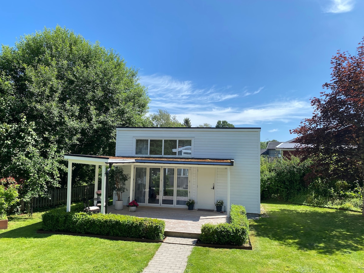 A modern cottage is surrounded by a lush garden, featuring a spacious front deck with a pergola. Large glass doors provide natural light, while a neatly landscaped lawn enhances the inviting outdoor area. The structure has a clean design, complemented by verdant greenery.