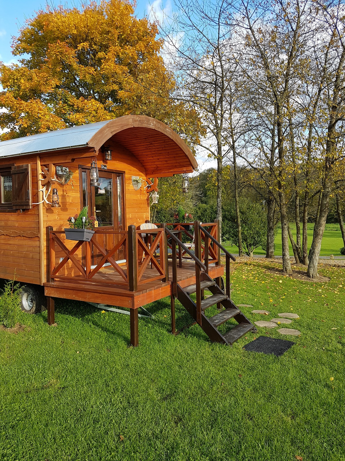 A charming wooden roulotte is situated on a grassy area, surrounded by trees with autumn foliage. Steps lead up to the entrance, which features a railing and potted plants. The warm wood exterior and curved roof create a welcoming appearance.