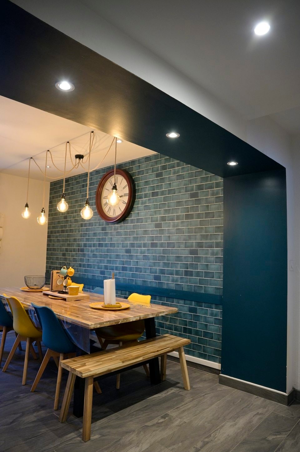 A dining area is presented with a wooden table and bench seating, accompanied by colorful chairs. A large wall clock and textured blue tile wall add character to the space. Pendant lights illuminate the area, highlighting the clean, modern design.