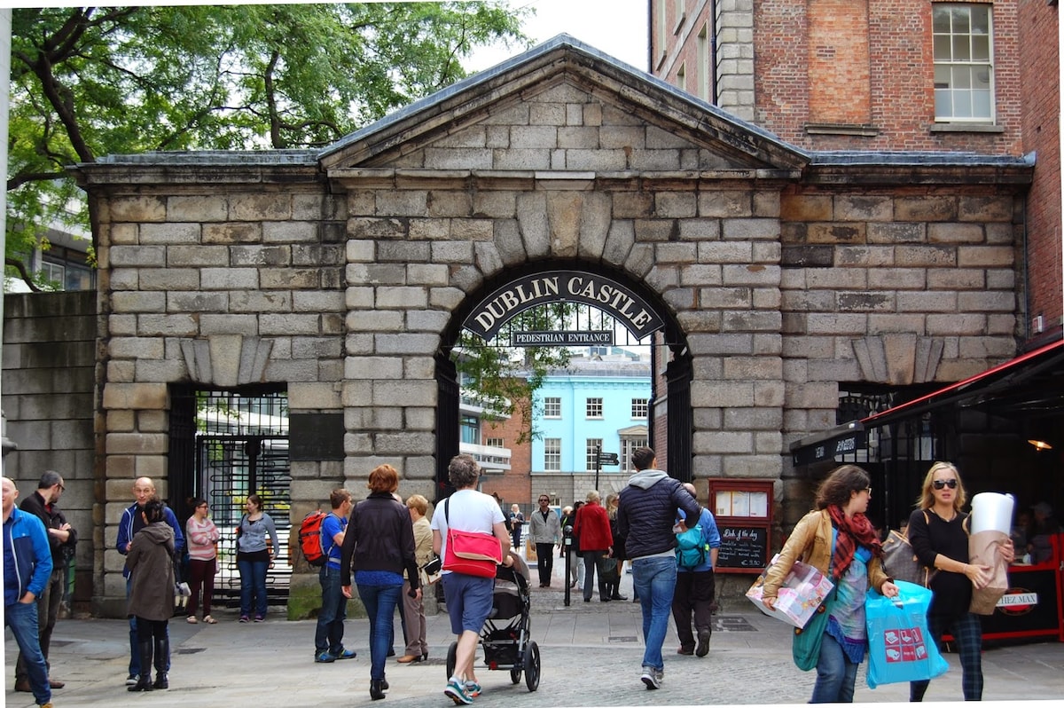 An arched stone entrance of Dublin Castle welcomes visitors through its grand doorway. People of varying ages and backgrounds stroll through the archway, engaged in conversation. The surrounding buildings show a mix of architectural styles, providing a glimpse of the vibrant atmosphere of Dublin's historic district.