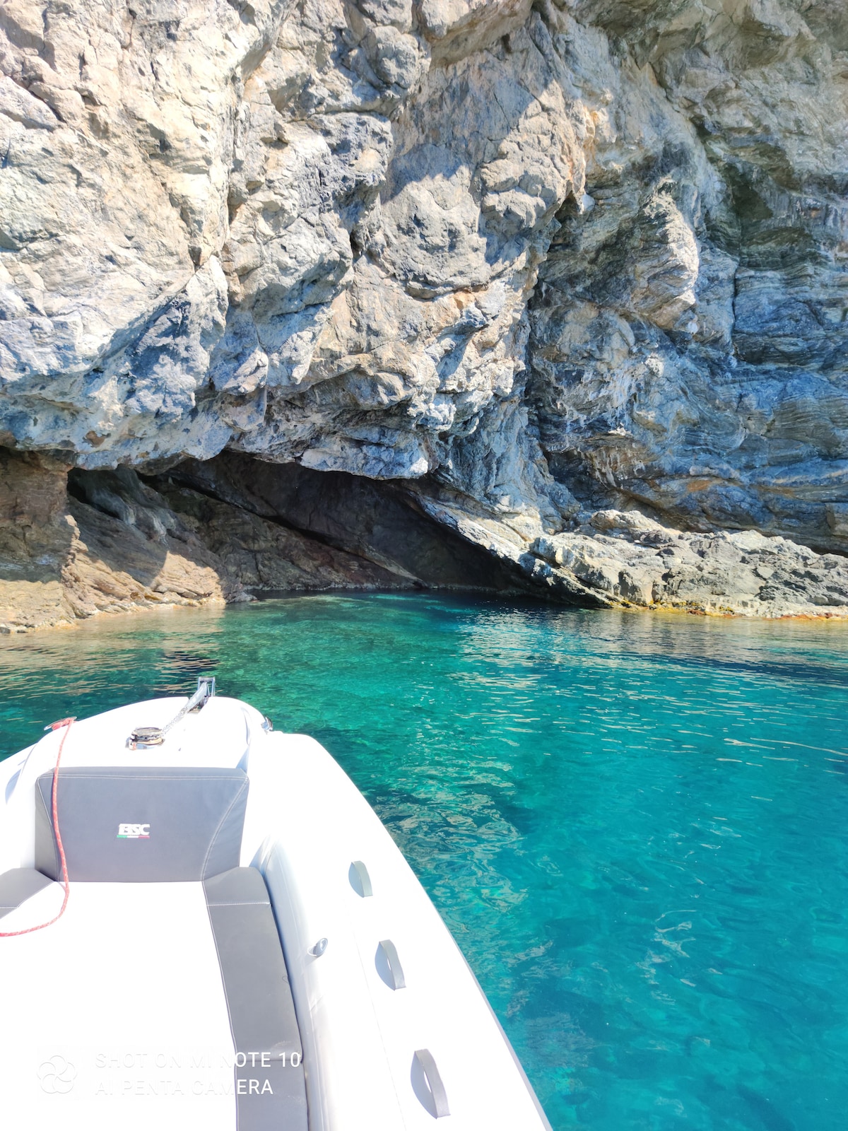 A motorboat is anchored near a rocky coastal area, where clear turquoise water meets jagged cliffs. The sun reflects off the water's surface, enhancing the vibrant colors of the surrounding nature.