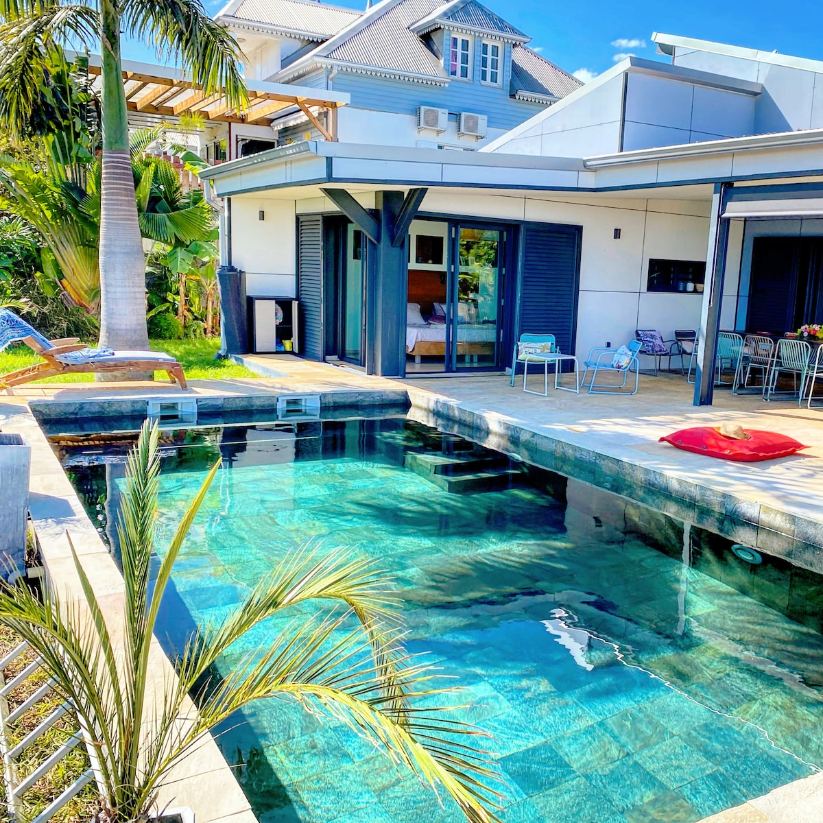 A modern villa is surrounded by lush greenery, featuring a glistening quartzite pool with clear blue water. Outdoor seating is available, complemented by wooden lounge chairs and a vibrant cushion. The design highlights a seamless flow between indoor and outdoor spaces.