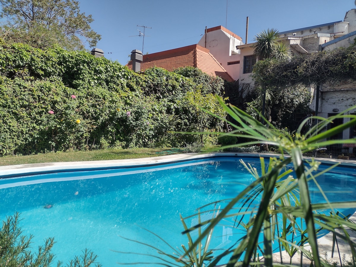 A refreshing pool is surrounded by lush greenery, featuring various plants and vibrant flowers. The clear water reflects the sunlight, creating a serene atmosphere. In the background, residential buildings are partially visible, adding context to the space.