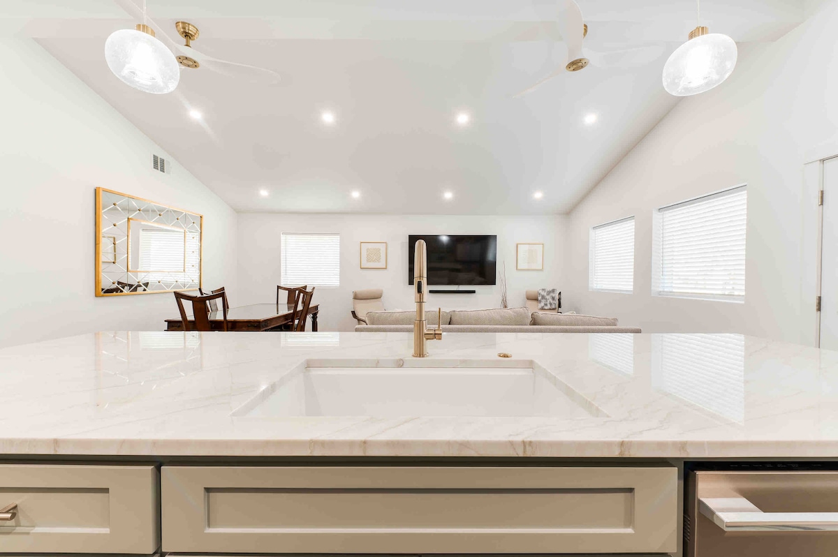 The image displays a modern open kitchen with a marble countertop and a spacious sink. A large window is visible in the background, alongside a comfortable living area featuring a sofa and a mounted television. Bright lighting enhances the neutral color scheme throughout the space.