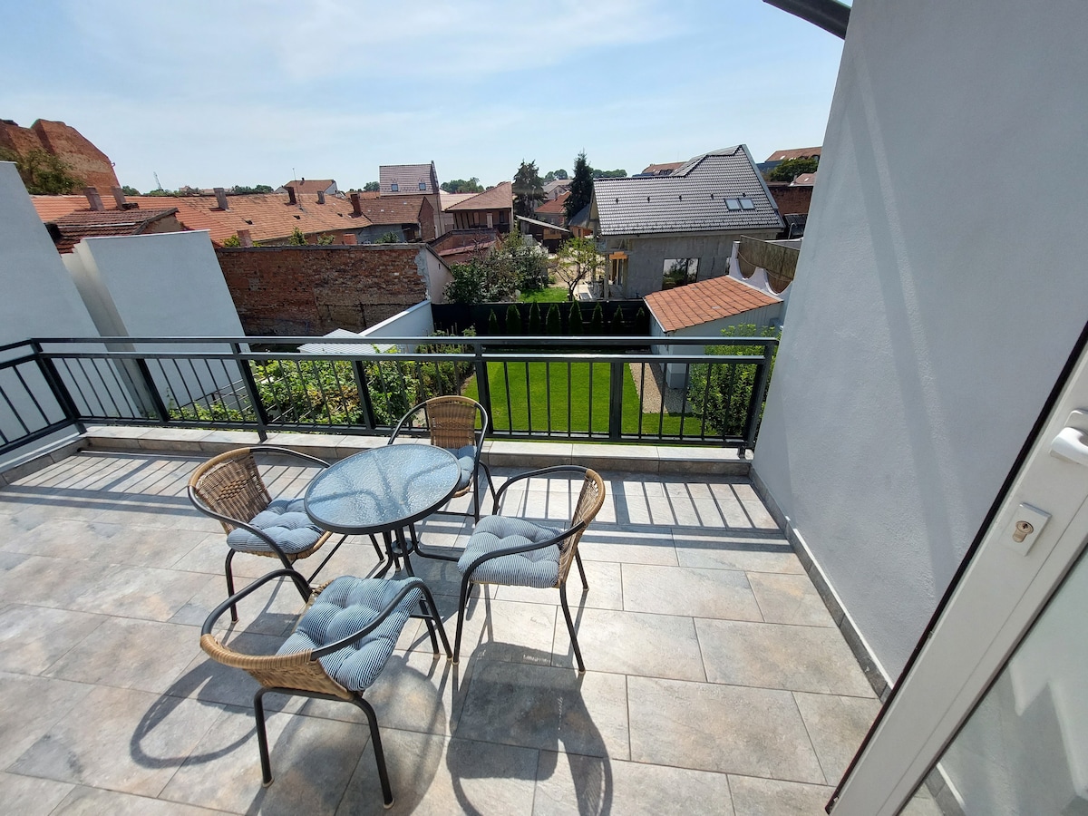 A spacious terrace is presented, featuring a round table surrounded by four wicker chairs. The area offers a view of a well-maintained garden and neighboring rooftops, illuminated by natural light. The terrace's railing enhances the open feeling of the outdoor space.