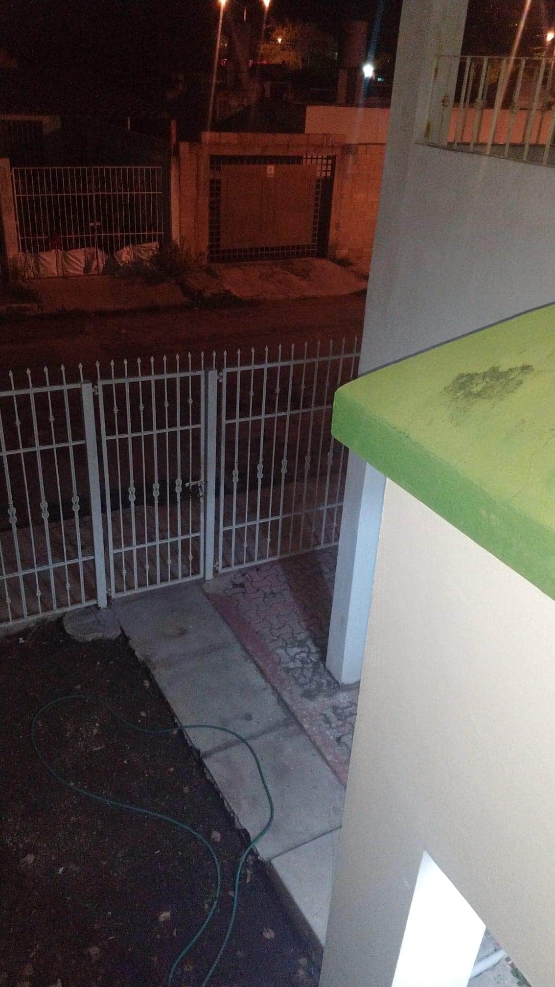 A view from a second-floor angle shows a private parking area bordered by white metal gates. The ground reveals a tiled pathway, with green and gray elements contrasting with the night sky. Soft lighting from nearby street lamps illuminates the surroundings.
