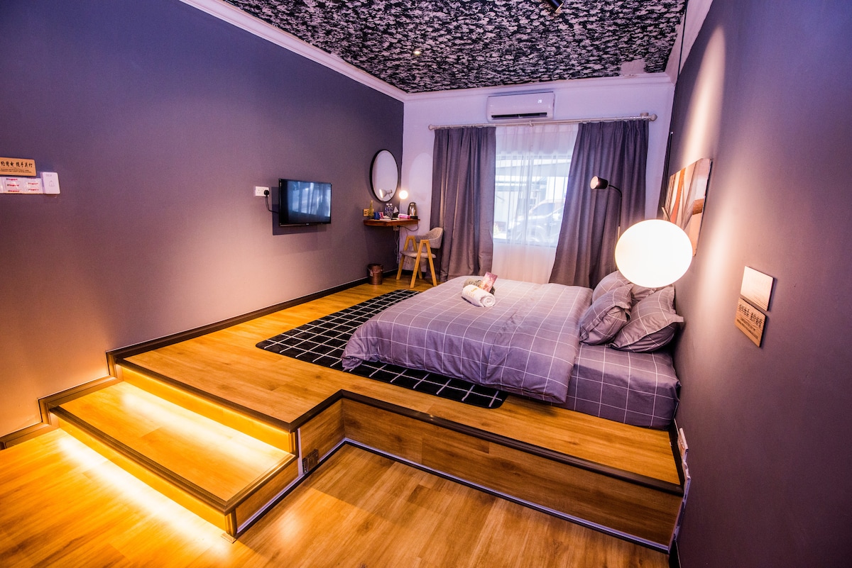 A modern bedroom features a spacious bed with neutral bedding, complemented by a soft area rug. Step-up wooden platforms lead into the room illuminated by soft lighting. A window with sheer curtains allows natural light, while a small desk area and a wall-mounted TV add functionality.