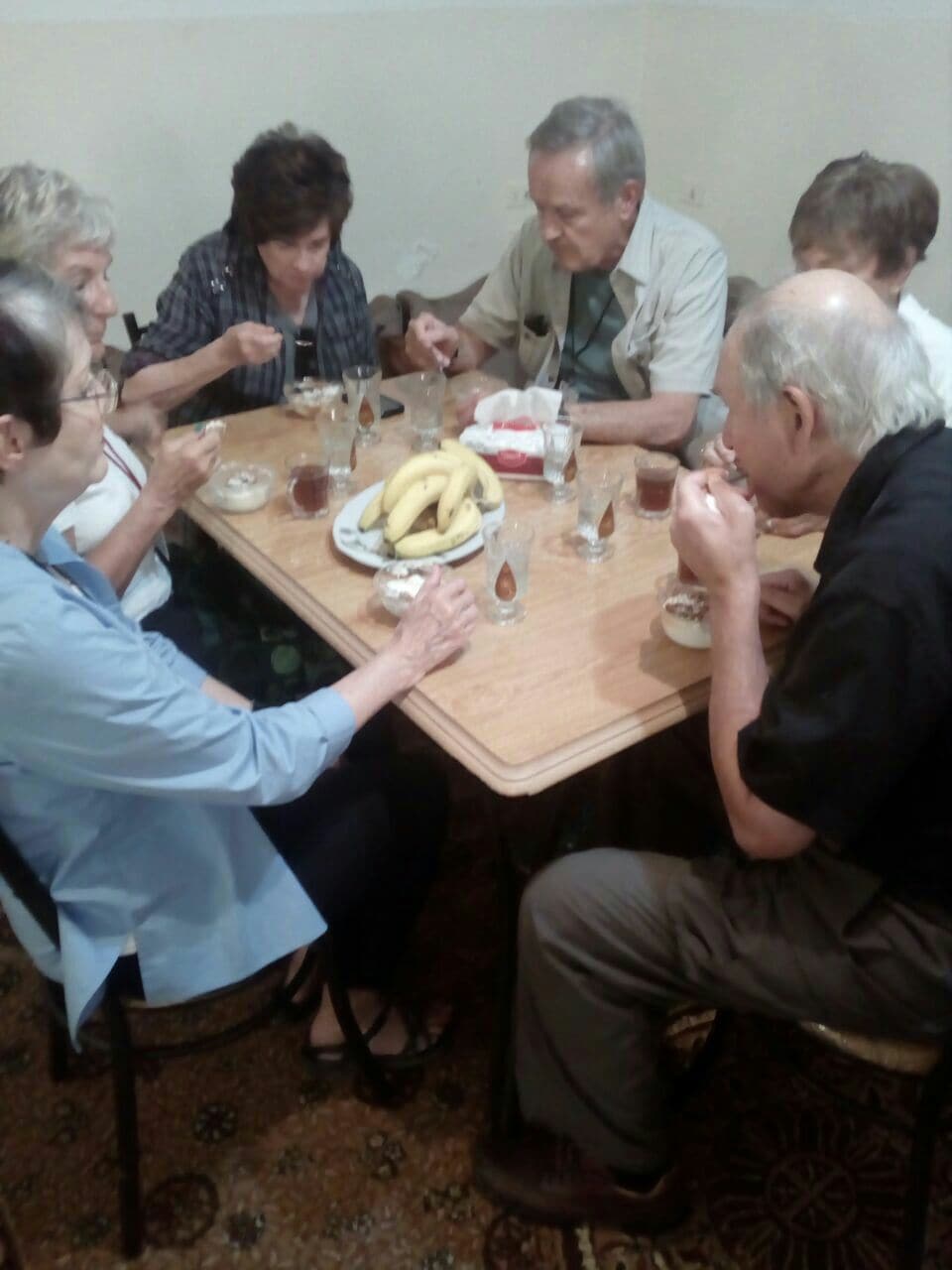 A wooden dining table is occupied by several individuals engaged in a meal. Bowls and glasses are visible on the table, and a plate of bananas is centered among other items. The room features simple decor with a patterned carpet beneath the chairs.