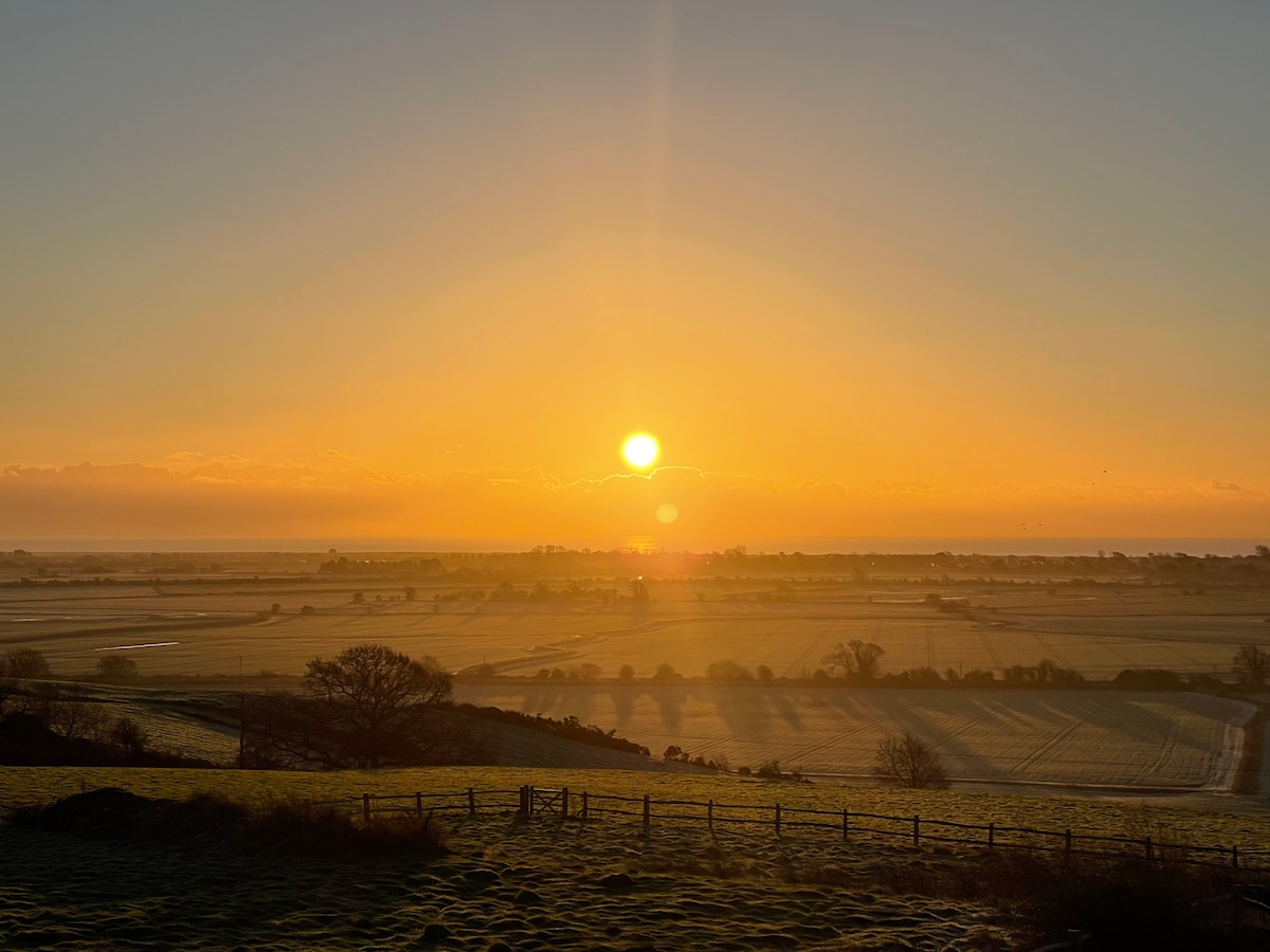 A scenic view captures a sunrise over expansive fields, with the soft glow of the sun rising on the horizon. The foreground features rolling hills and a wooden fence, while the shimmering blue of the sea is visible in the distance.