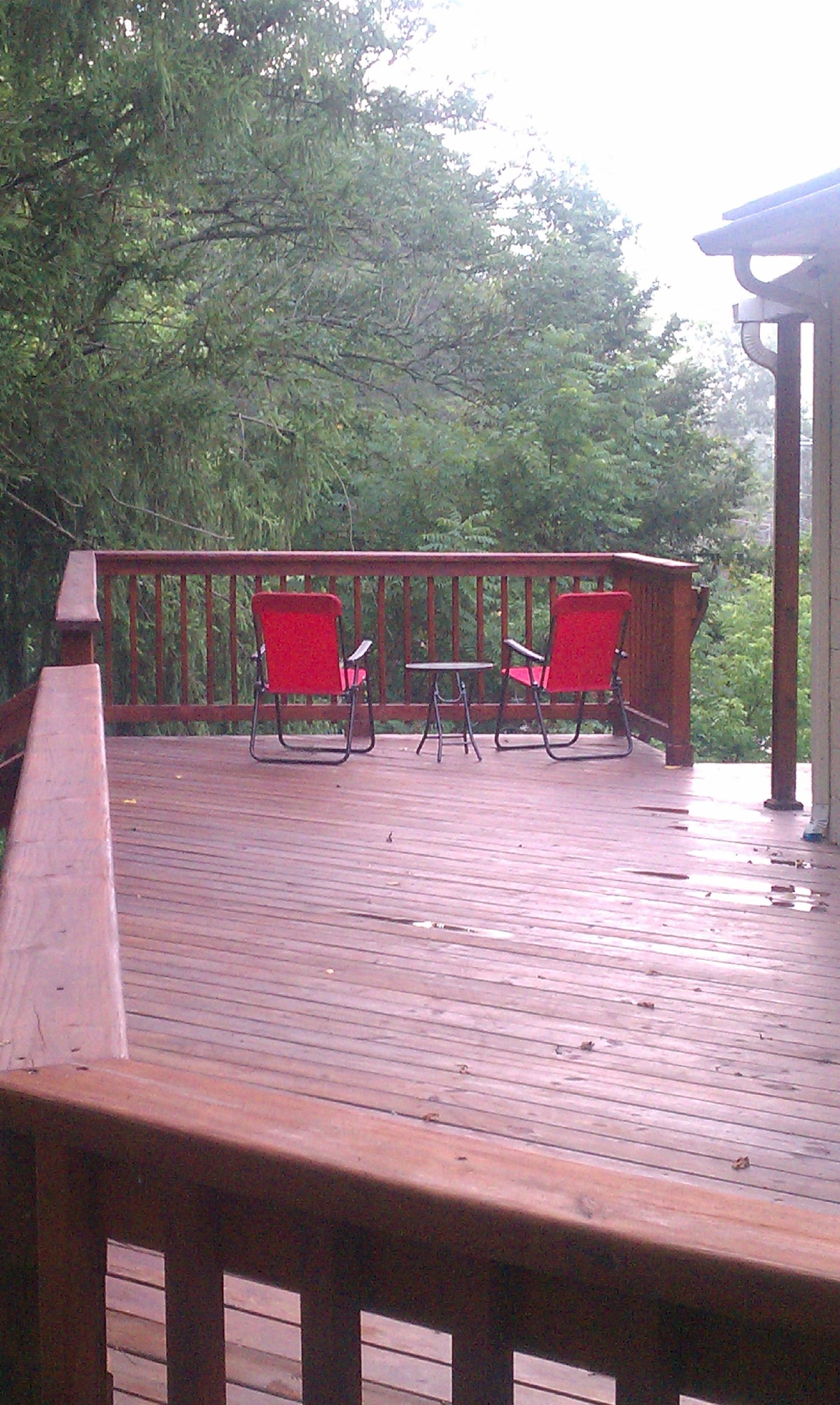 A spacious wooden deck is visible, featuring two red chairs and a small black table. The surrounding greenery enhances the setting, providing a peaceful outdoor space for relaxation. Natural light filters through the trees, creating a welcoming atmosphere.