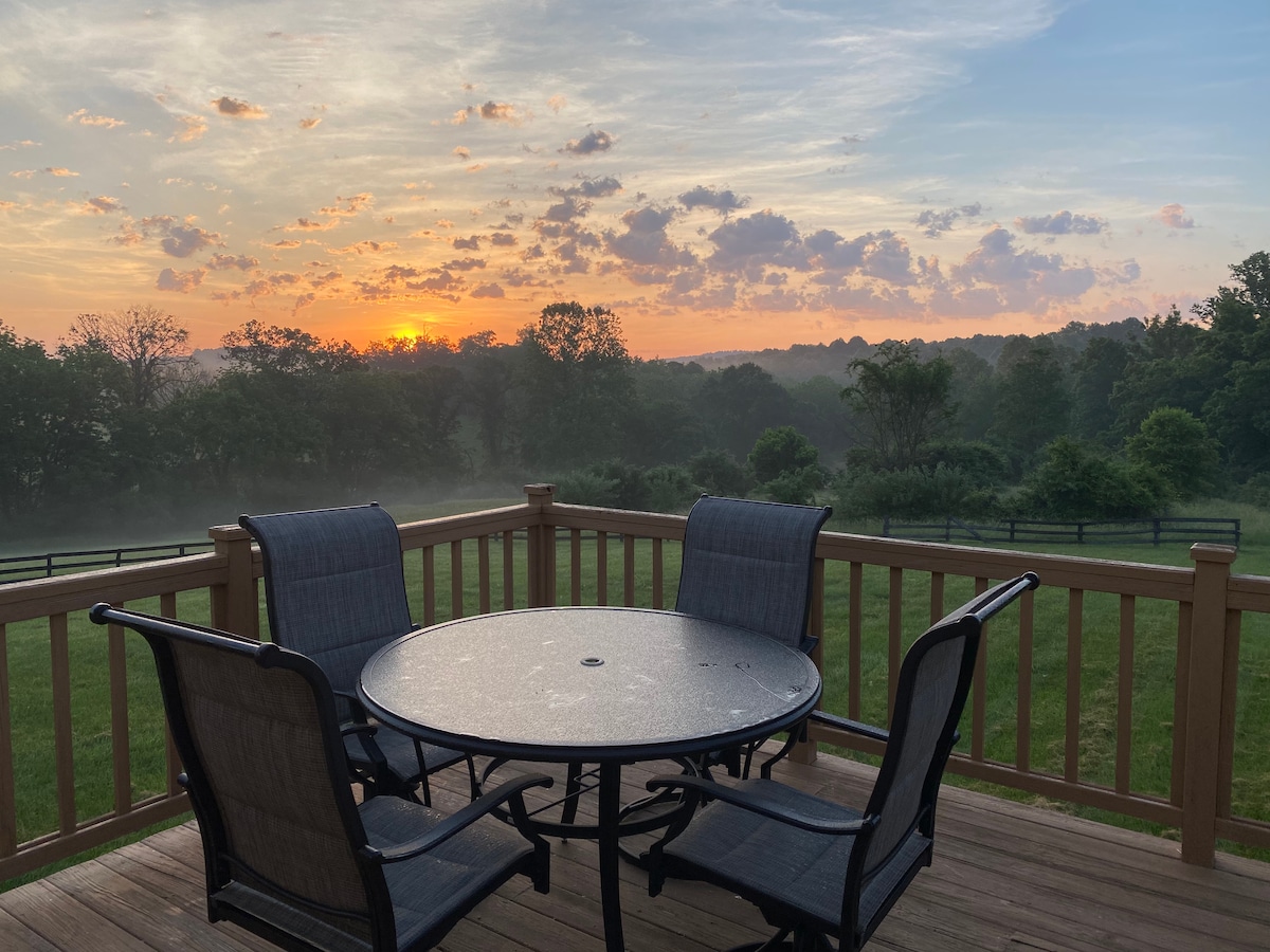 A wooden deck overlooks a misty landscape at sunrise, featuring a round table surrounded by four chairs. Soft hues of orange and pink fill the sky, while trees are silhouetted against the light in the background, creating a serene setting.