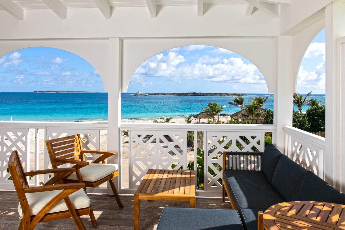 A spacious outdoor seating area is visible, featuring wooden chairs and a table, with a protected view of calm ocean waters and a sandy beach. Arched openings allow natural light to brighten the space, while the surrounding vibrant greenery adds a touch of nature.