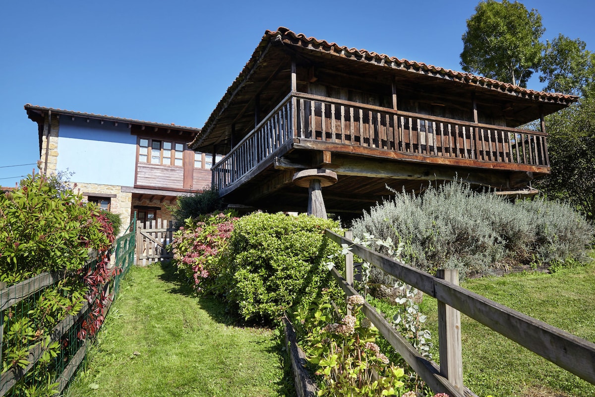 A renovated cottage is presented with a raised wooden terrace supported by sturdy posts. Lush greenery and colorful flowers surround the property, with a clear blue sky above. The structure features both wooden siding and stone accents, enhancing its rustic charm.