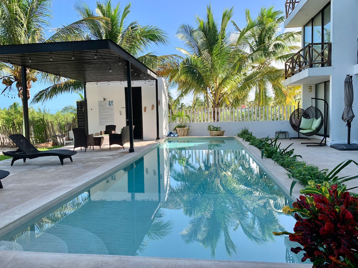 The image depicts a serene outdoor area featuring a calm swimming pool surrounded by lush greenery and palm trees. A shaded seating area with modern chairs is visible, enhancing the tranquil space. The clear blue sky complements the inviting atmosphere.