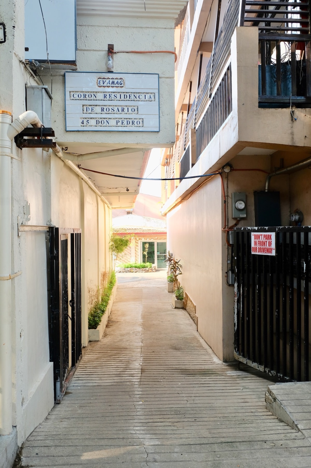 A narrow pathway leading to the entrance of Coron Residencia de Rosario is flanked by beige walls. Potted plants line the corridor, while a sign for the residence hangs above. The ground is paved with wooden slats, providing a clear route towards the welcoming entrance.