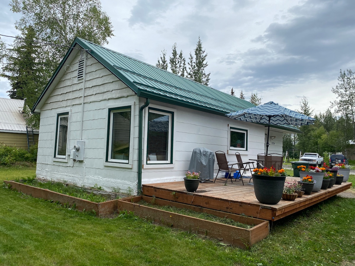 The exterior of a charming two-bedroom home is depicted, featuring a deck adorned with potted flowers. A blue umbrella provides shade over outdoor seating, with a grassy area surrounding the property. Large windows allow for ample natural light, complemented by a green metal roof.