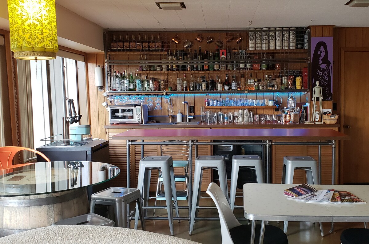 A well-organized bar area features a wooden countertop with a variety of glassware and bottles displayed on shelves above. Seating for four is provided with metallic stools. A round table with a metal base and a light fixture overhead completes the space, illuminated by natural light from windows.
