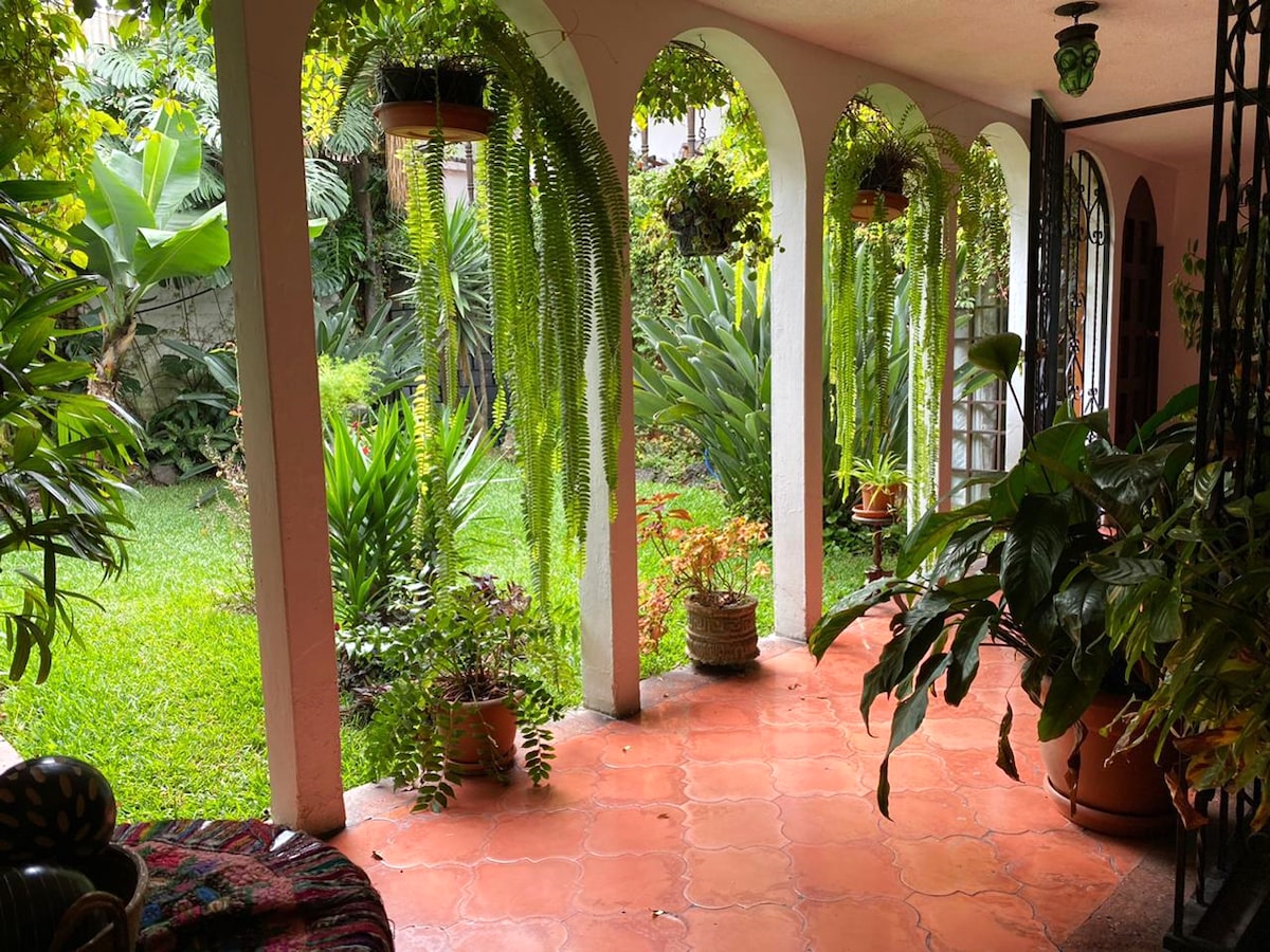 A covered walkway is framed by arched columns, leading into a lush garden. Green plants hang from the ceiling, while various tropical plants and flowers fill the space, providing a vibrant atmosphere. The terracotta flooring complements the natural greenery.
