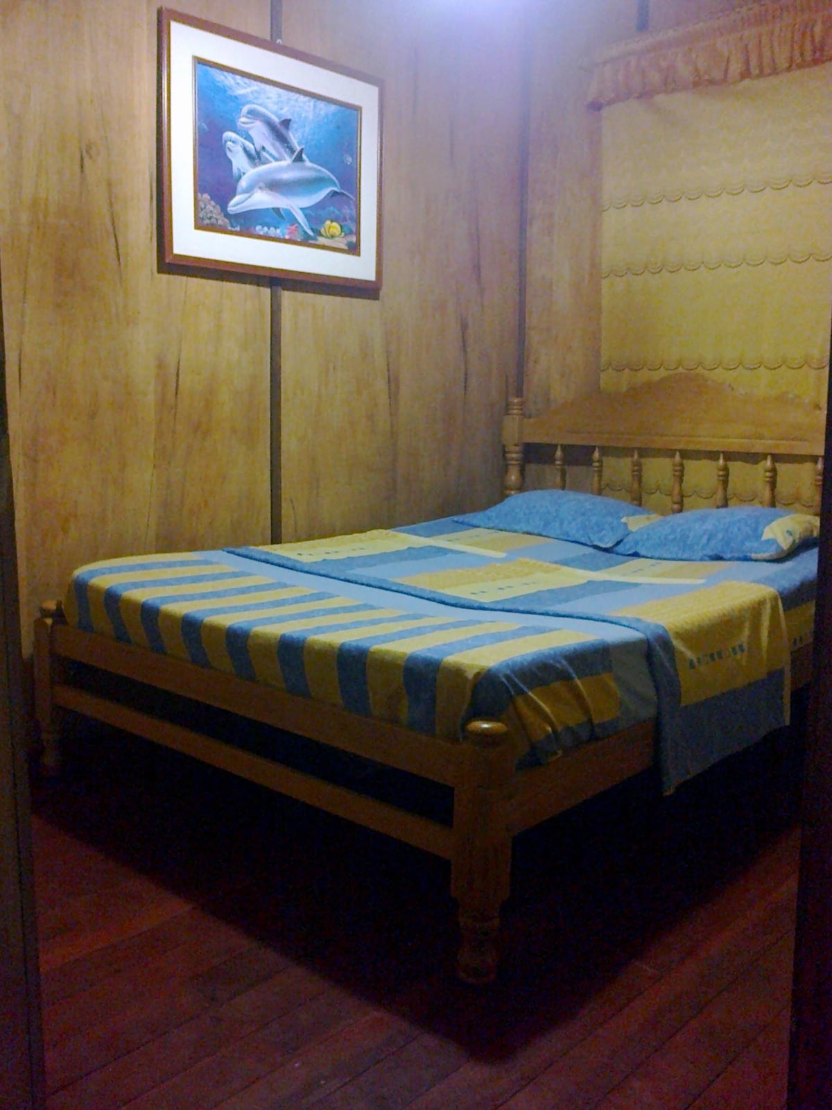 A small sleeping area features a wooden bed dressed in a blue and yellow striped blanket. Two light blue pillows rest on the bed, and a framed artwork depicting marine life hangs on the wall. Natural wood paneling creates a warm, inviting space.