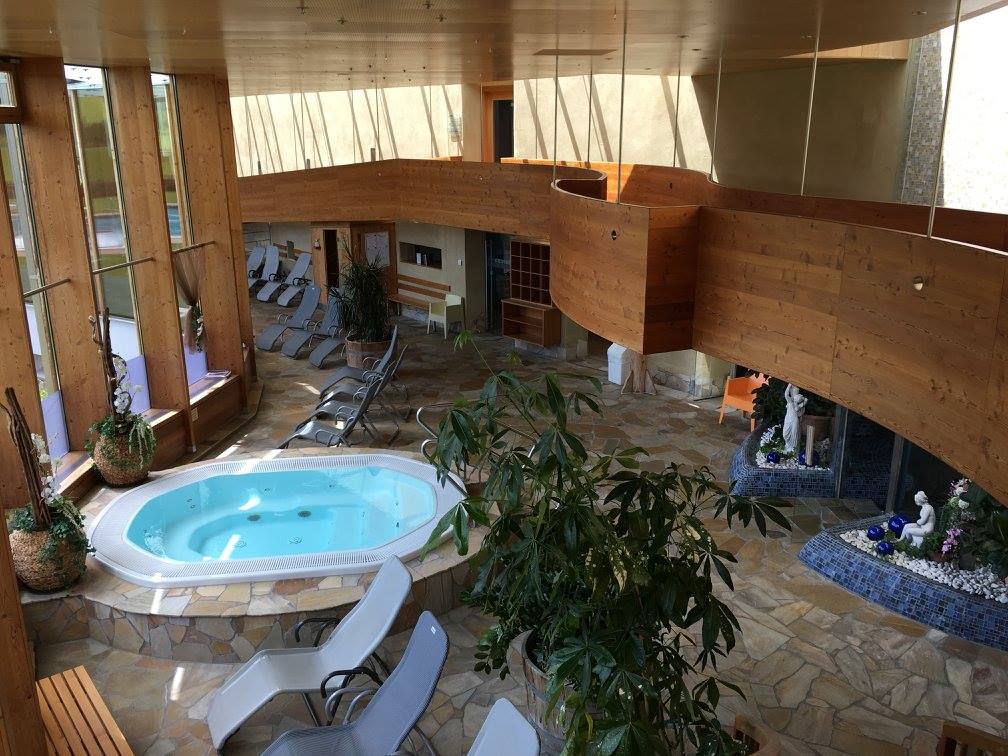 An indoor wellness area features a hot tub surrounded by comfortable loungers. Natural light floods the space through large windows, illuminating the wooden architectural details and stone flooring. Plants are positioned throughout, adding a touch of greenery to the serene atmosphere.
