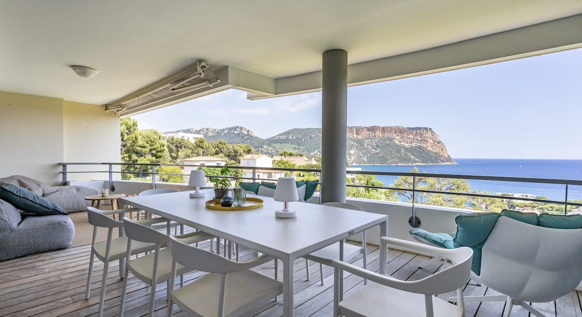 An outdoor terrace features a large dining table surrounded by several chairs, with soft lounge seating in the corners. The stunning view showcases the sea and the cliffs, providing a serene backdrop for gatherings.