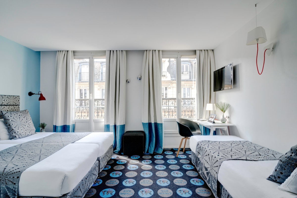 A bright hotel room features three individual beds arranged with patterned bedding, and large windows that provide natural light. A small desk is positioned near the window, complemented by a chair. A striking area rug adds color to the room's design.