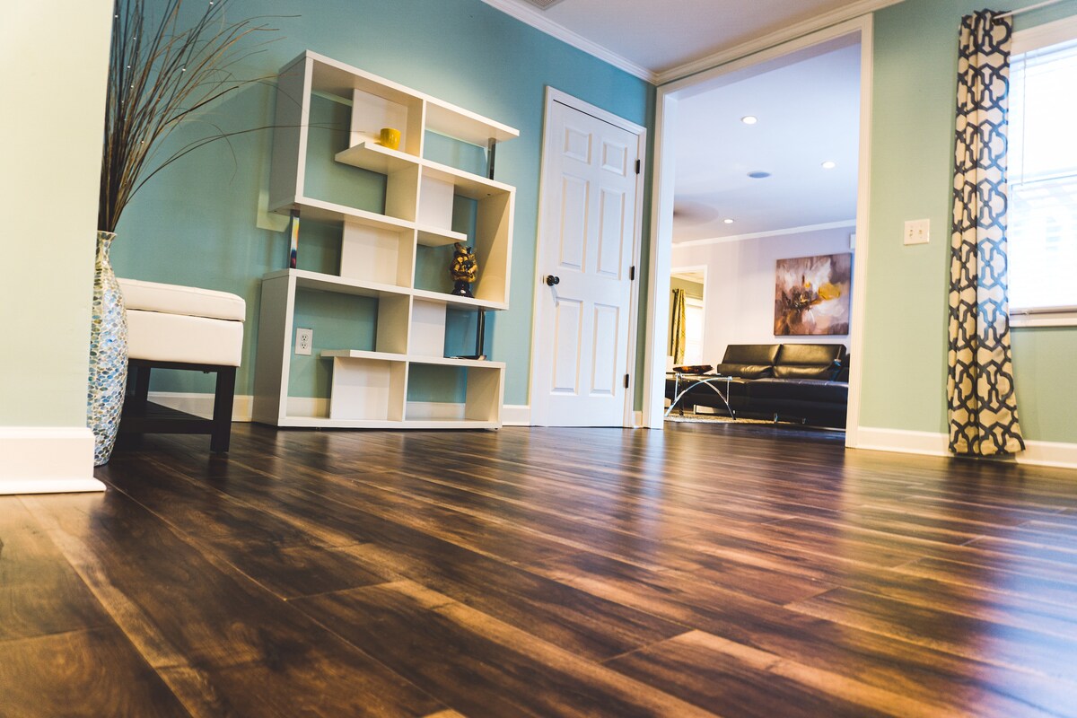 The image showcases a spacious room with hardwood flooring, accented by light teal walls. A white shelving unit stands against the wall, complemented by a decorative vase. An open doorway leads to a separate area, while a modern seating arrangement is visible in the background.