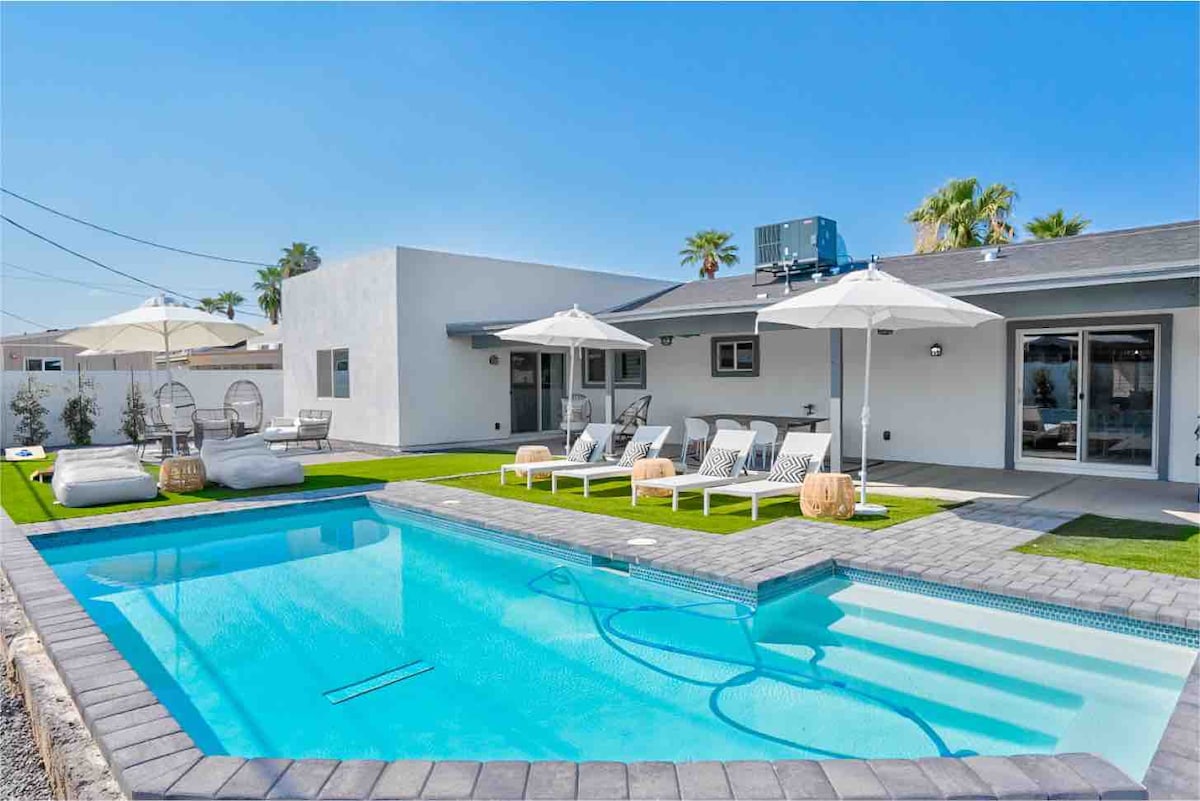 An outdoor space showcases a heated pool surrounded by lounge chairs and umbrellas. Modern seating is arranged on the grass, offering a place to relax. The patio leads to the house’s large glass doors, providing access to the indoor living area.